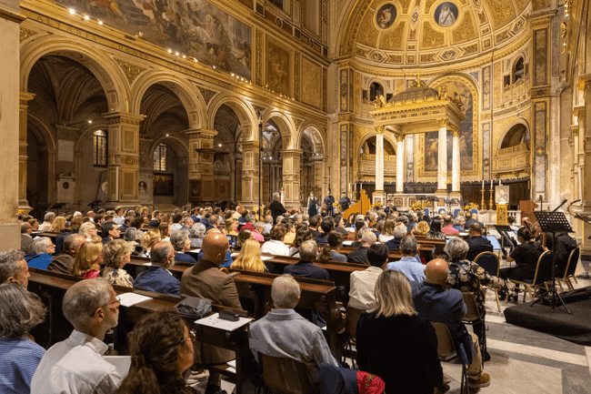 Panoramica della chiesa affrescata e piena di spettatori, con il coro e l’orchestra che si preparano all’esecuzione sul presbiterio.