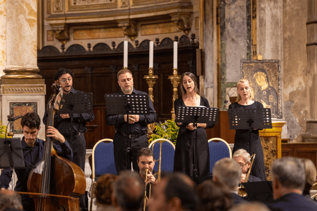 Quattro cantanti, due uomini e due donne, eseguono un brano di musica sacra accompagnati da un ensemble strumentale, sullo sfondo l’altare riccamente decorato.