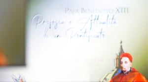 Dettaglio sfocato del titolo della conferenza “Papa Benedetto XIII: Profezia e Attualità di un Pontificato”, con un’illustrazione del pontefice in primo piano.