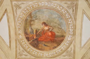 Affresco rinascimentale circolare con una donna seduta che spegne un fuoco con un recipiente d’acqua, all’interno di una cornice decorata con motivi floreali