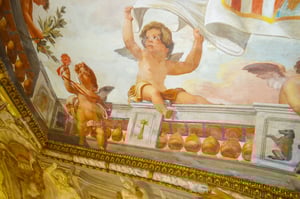 Dettaglio di un affresco barocco con due putti alati: uno seduto sul parapetto mentre regge un drappo bianco, l’altro in piedi che solleva un fiore rosso. Colori vivaci e cornice dorata decorano la scena