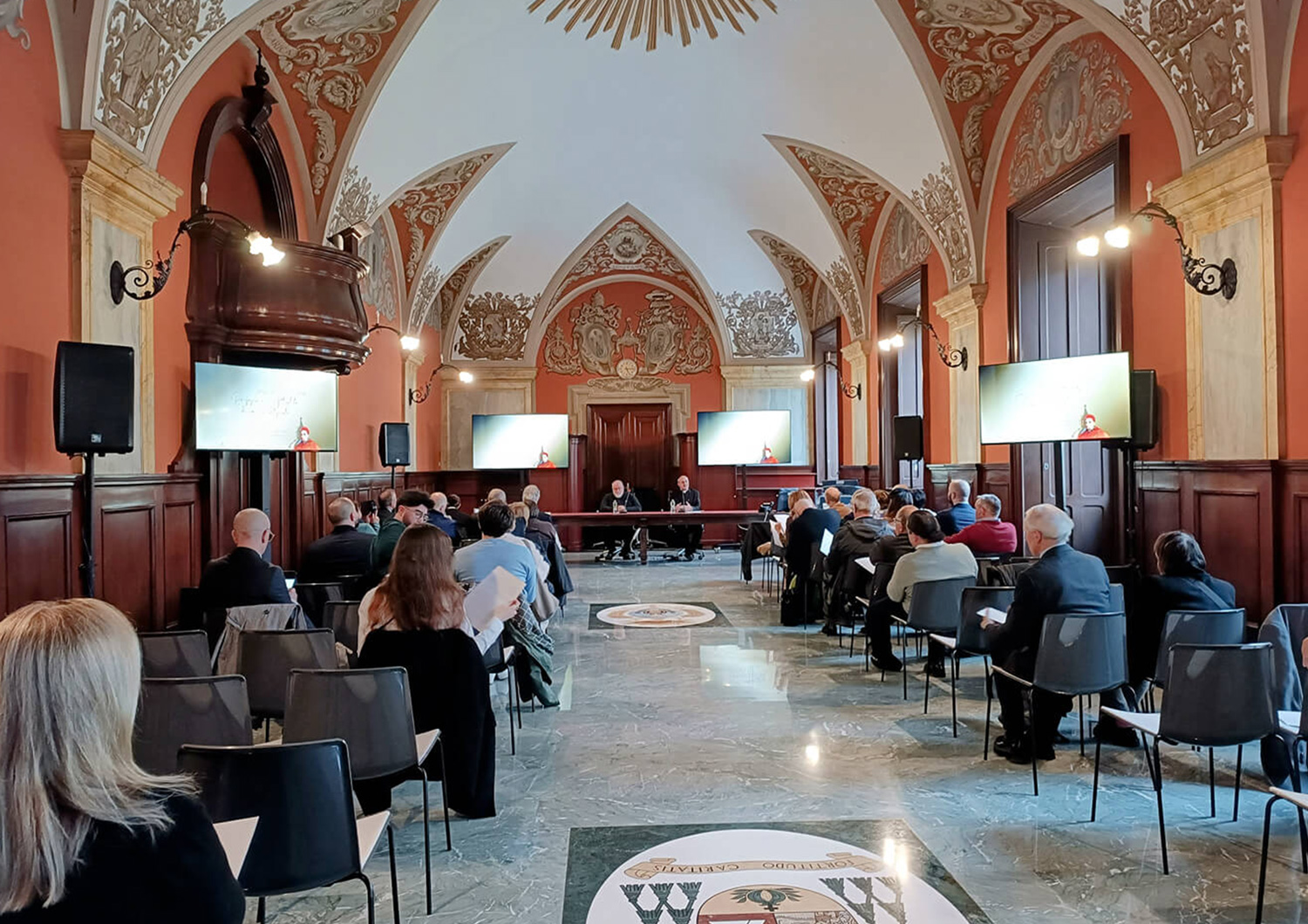 Sala conferenze storica con soffitti affrescati e pareti decorate, dove un gruppo di persone è seduto ad assistere a un incontro o convegno; due relatori sono seduti a un tavolo sul palco, con schermi laterali che proiettano immagini Sala conferenze storica con soffitti affrescati e pareti decorate, dove un gruppo di persone è seduto ad assistere a un incontro o convegno; due relatori sono seduti a un tavolo sul palco, con schermi laterali che proiettano immagini