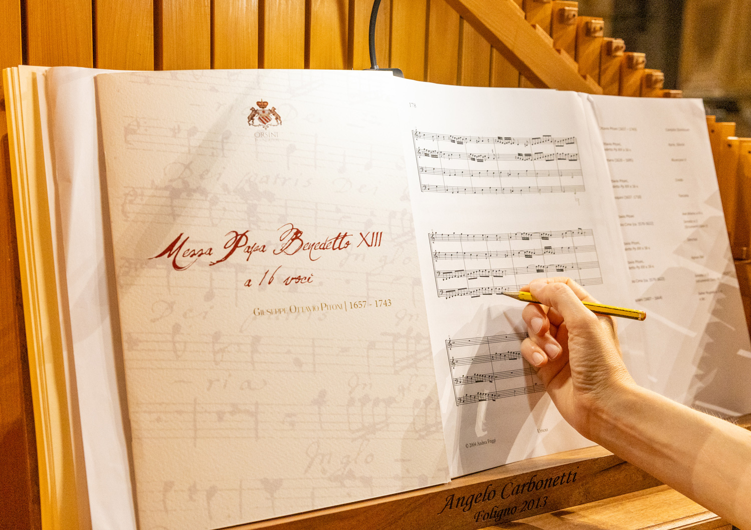 Primo piano di uno spartito aperto sul leggio di un organo, con il titolo ‘Messa Papa Benedetto XIII a 16 voci’ di Giuseppe Ottavio Pitoni; una mano tiene una matita puntata sulle note musicali Primo piano di uno spartito aperto sul leggio di un organo, con il titolo ‘Messa Papa Benedetto XIII a 16 voci’ di Giuseppe Ottavio Pitoni; una mano tiene una matita puntata sulle note musicali